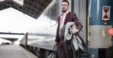 stylish man standing near train with bag and coat in railway station