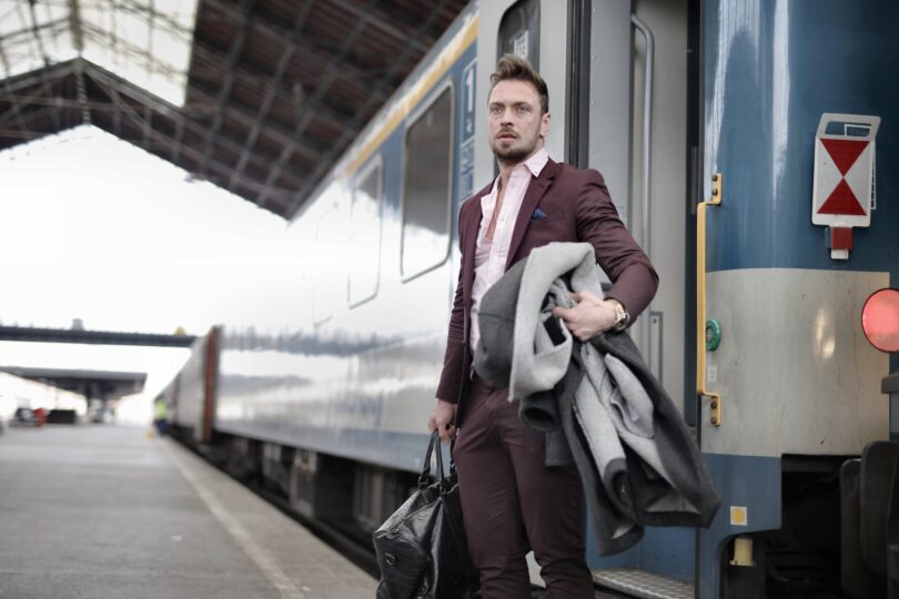 stylish man standing near train with bag and coat in railway station