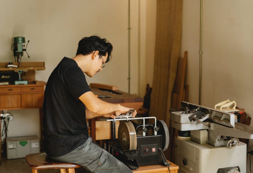 attentive ethnic artisan sharpening tool on grinding machine in workroom
