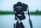 selective focus photo of black canon camera on tripod stand in front of body of water photo