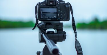selective focus photo of black canon camera on tripod stand in front of body of water photo